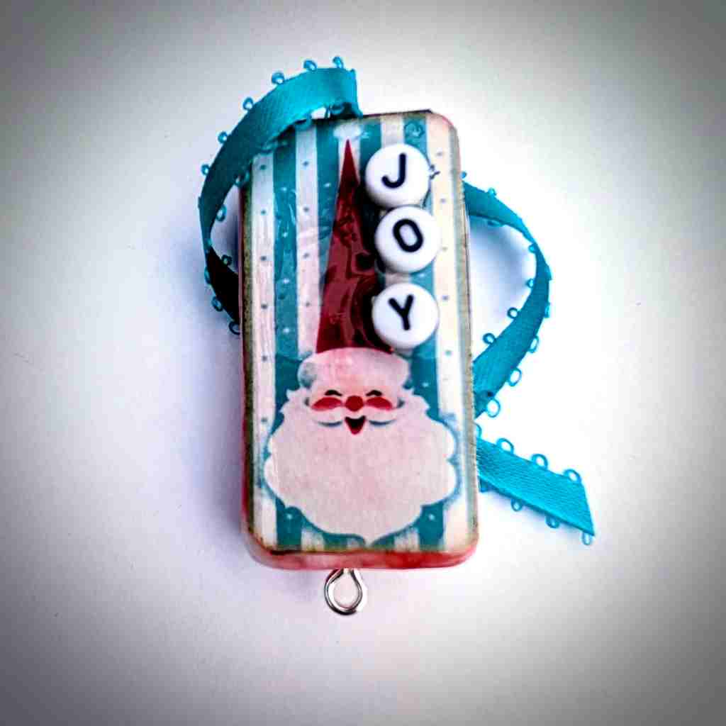 Decorative ornament with Santa Claus design and 'JOY' text in beads made of a domino on a light gray background