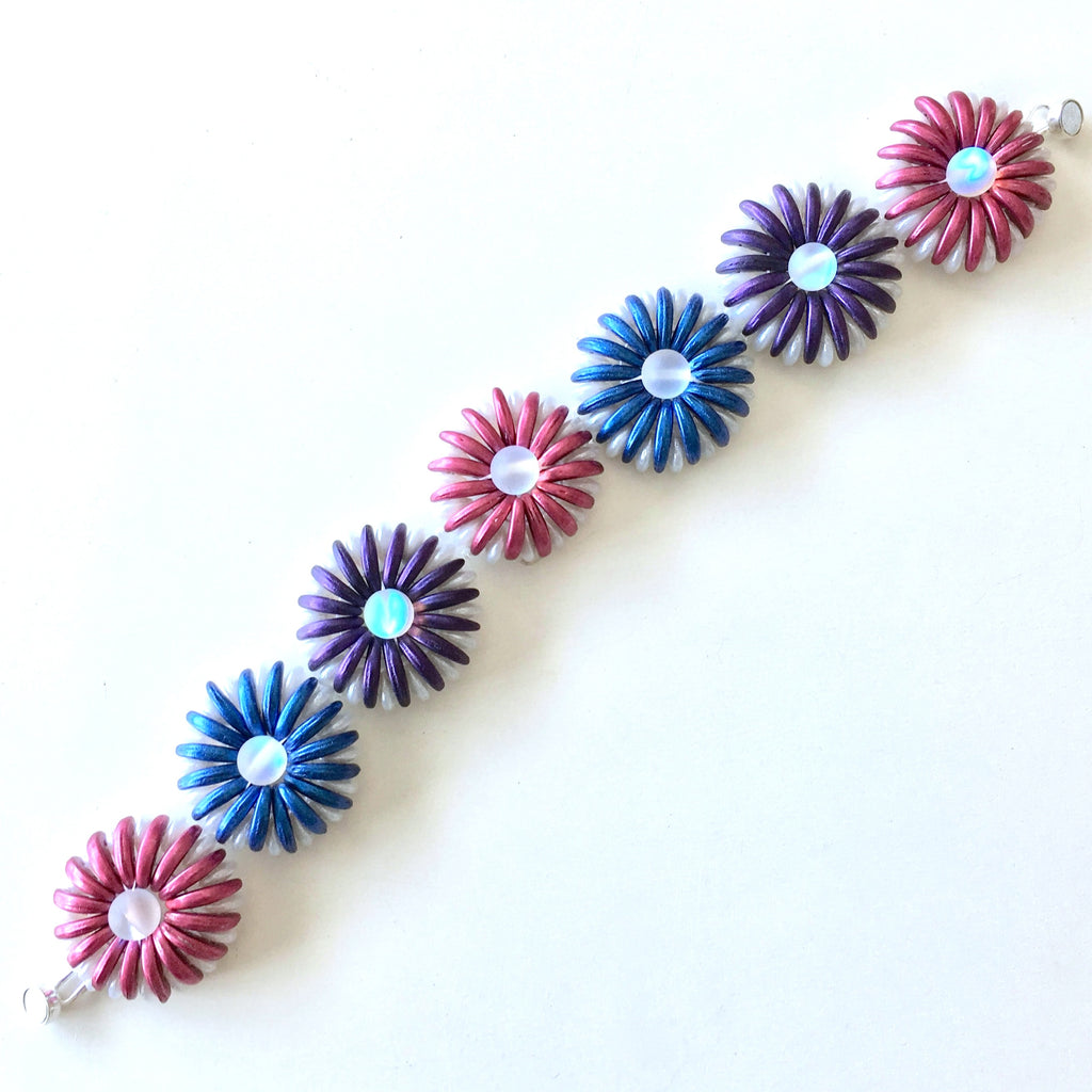A handmade beaded bracelet featuring alternating flower-shaped links in shades of blue, purple, and pink, each with a small iridescent light blue center, displayed on a clean white background.
