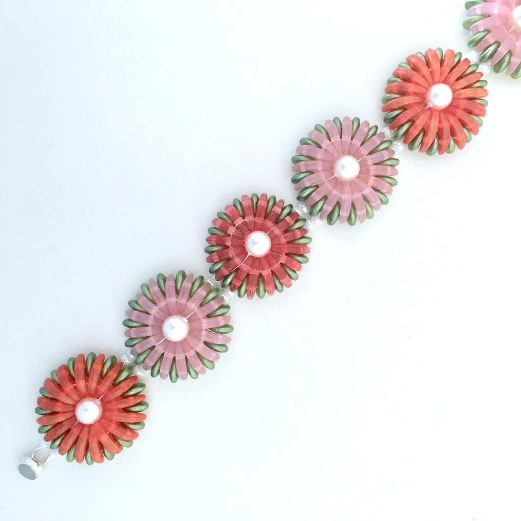 A close-up of a floral bracelet featuring alternating pink and coral-red flower petals with green edges and white pearl-like centers, laid diagonally on a plain white background.