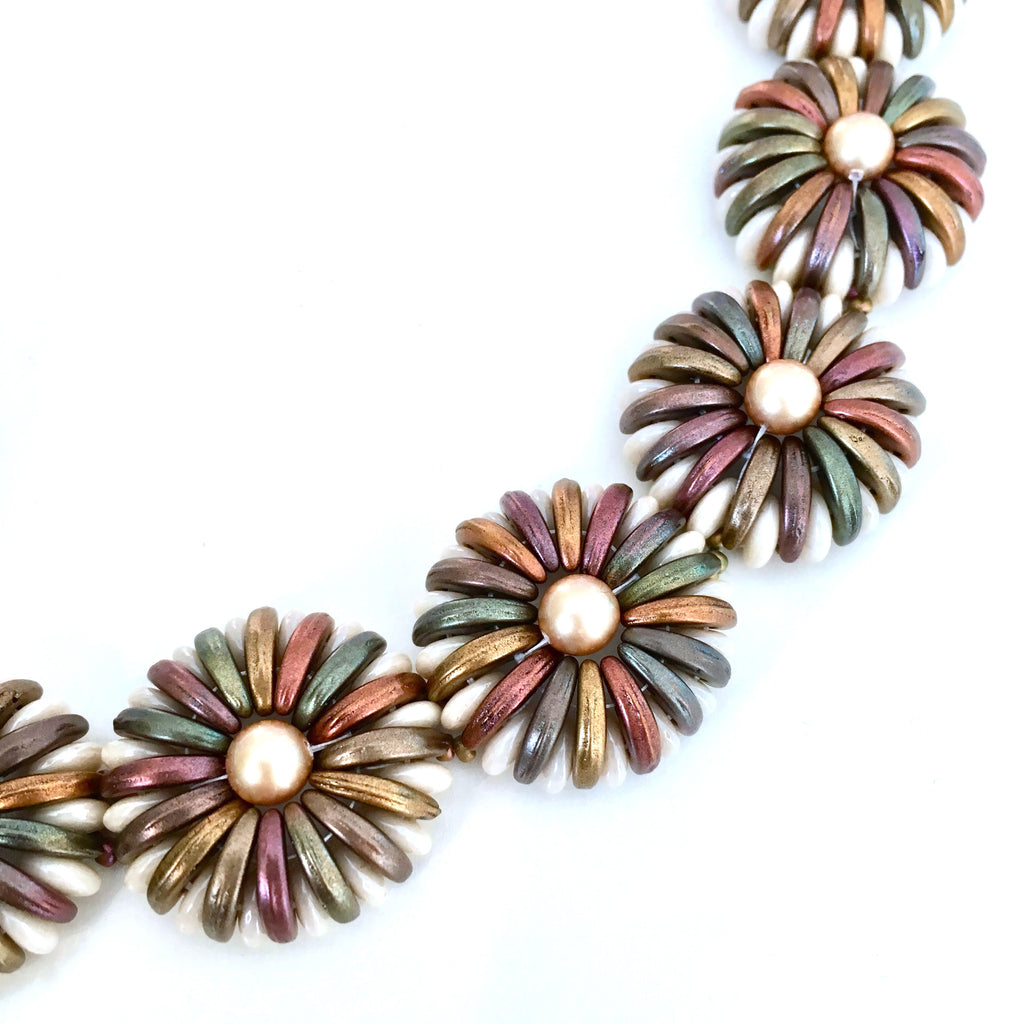 A close-up of a beaded bracelet featuring metallic flower-shaped beads with bronze, green, and pink patina rays surrounding a central cream pearl, set against a plain white background.