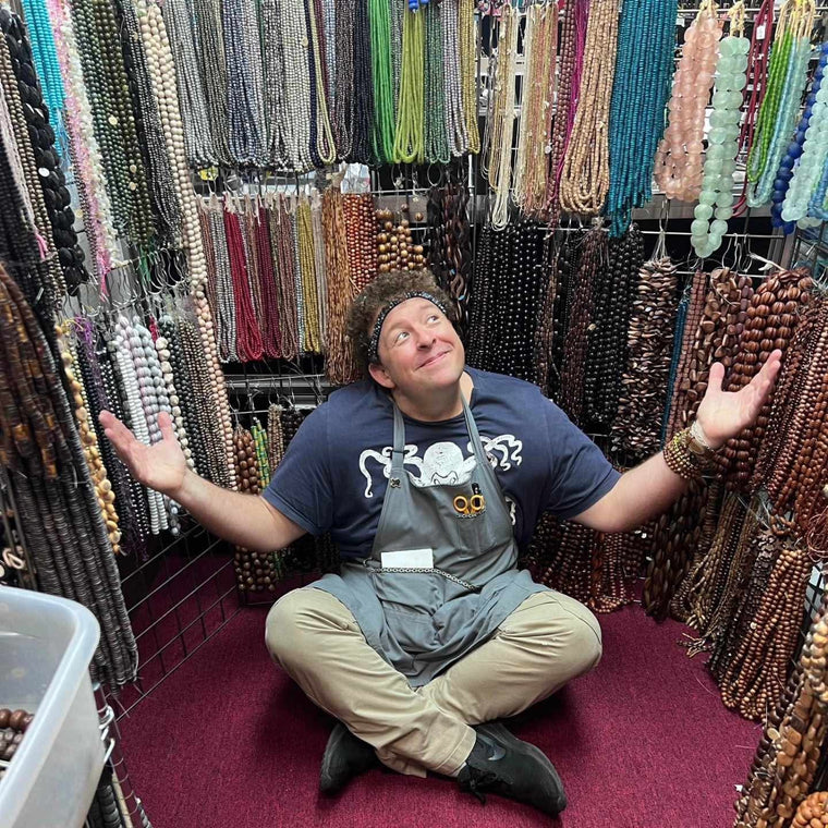 A person with a cheerful expression sitting amidst a variety of beads and jewelry in a store.