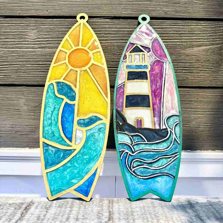 Two wooden surfboard-shaped panels with a sun and wave design made with resin, and a resin lighthouse and wave design, against a wooden background.